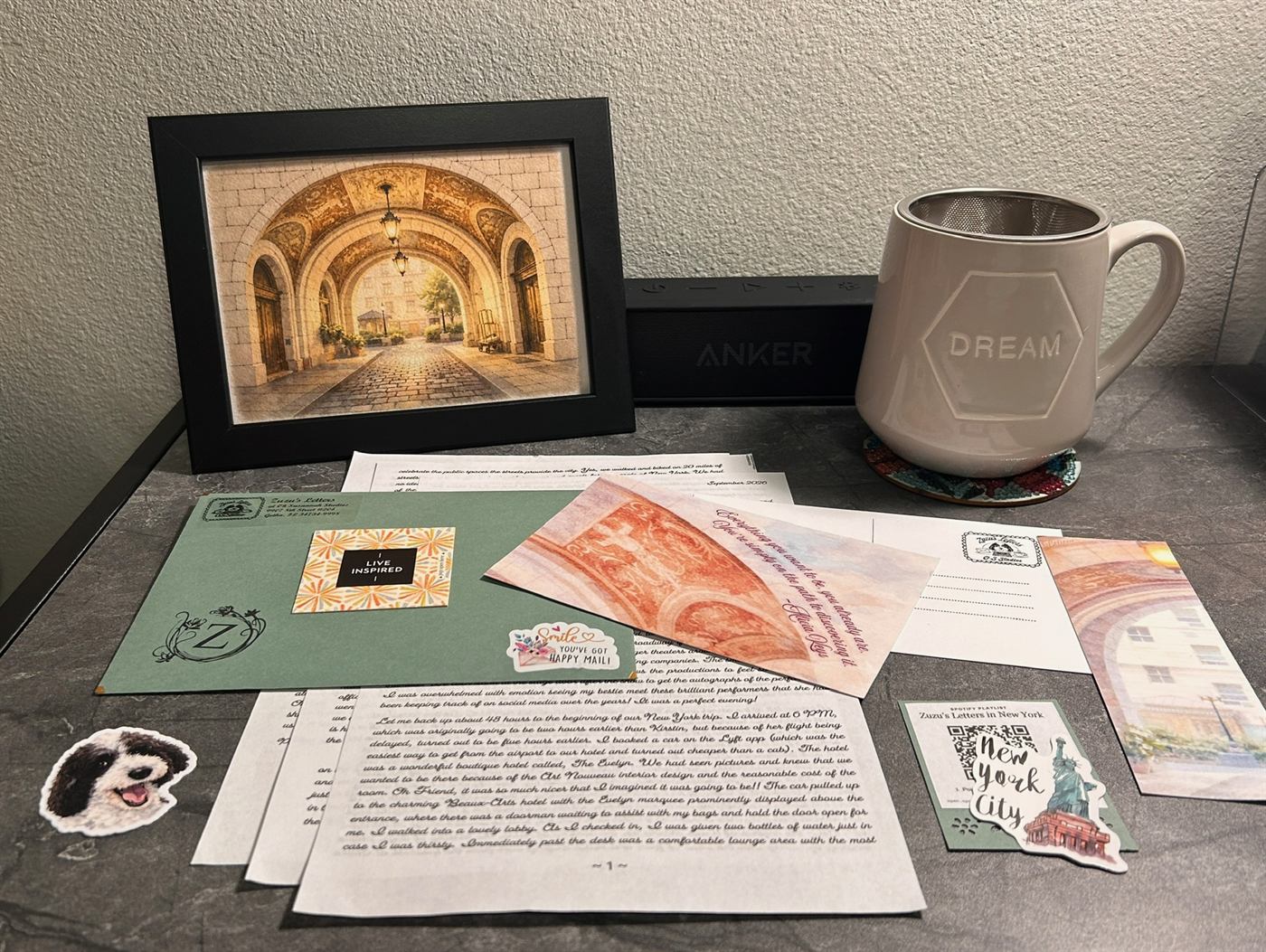 September&rsquo;s letter spread across a slate-grey table: the letter pages in Zuzu&rsquo;s script, the sage envelope with its stickers, the quote card, the bookmark and the playlist card, with the print of a sunlit stone archway framed in black standing behind them beside a mug.