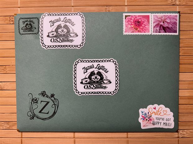 A sage green envelope on a bamboo mat, sealed and stamped: the Zuzu&rsquo;s Letters mark, a scrolled Z monogram, two pink dahlia Forever stamps, and a sticker reading Smile, you&rsquo;ve got happy mail.
