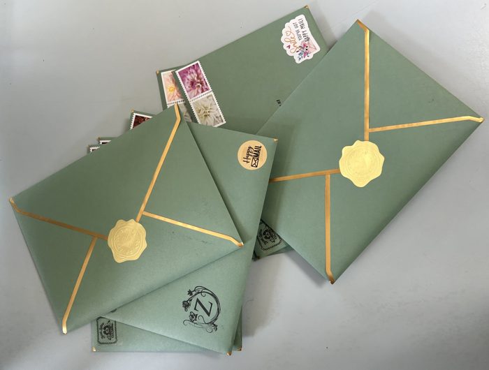 A fan of sage green Zuzu&rsquo;s Letters envelopes with gold-edged flaps, gold seals, flower stamps, and Happy Mail stickers.
