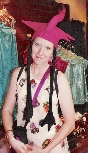 Susannah in a tall pink flamingo hat and an embroidered vest, standing in front of a rack of costumes.