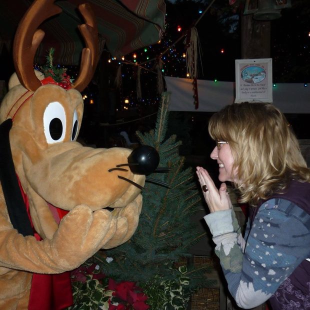 Zuzu clasping her hands in delight as Pluto, wearing reindeer antlers, poses beside a little Christmas tree under twinkling lights.