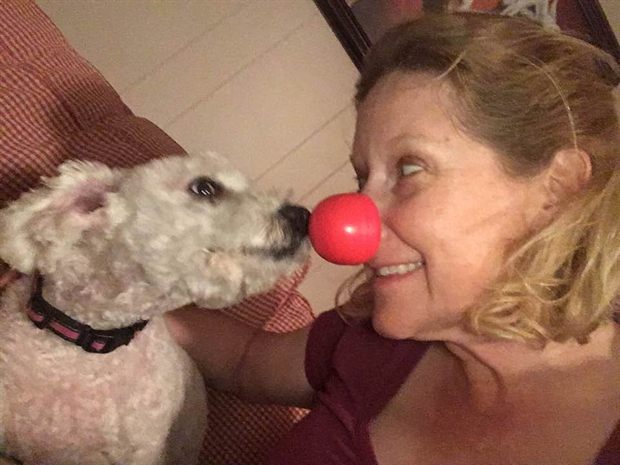 Susannah cheek to cheek with Gidget, her small white rescue dog, who is wearing a red clown nose.
