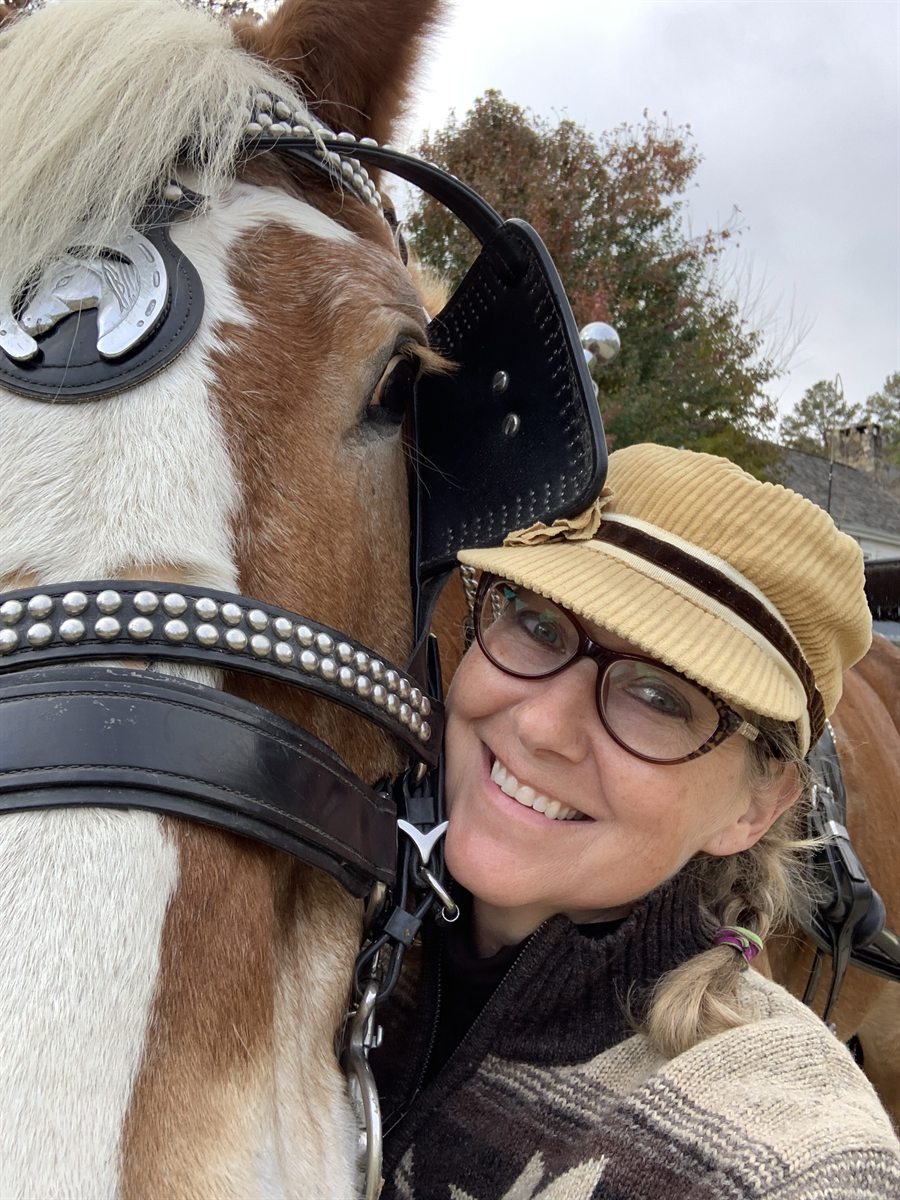 Zuzu in a corduroy cap and glasses, cheek to cheek with a chestnut and white horse in harness, both looking at the camera.